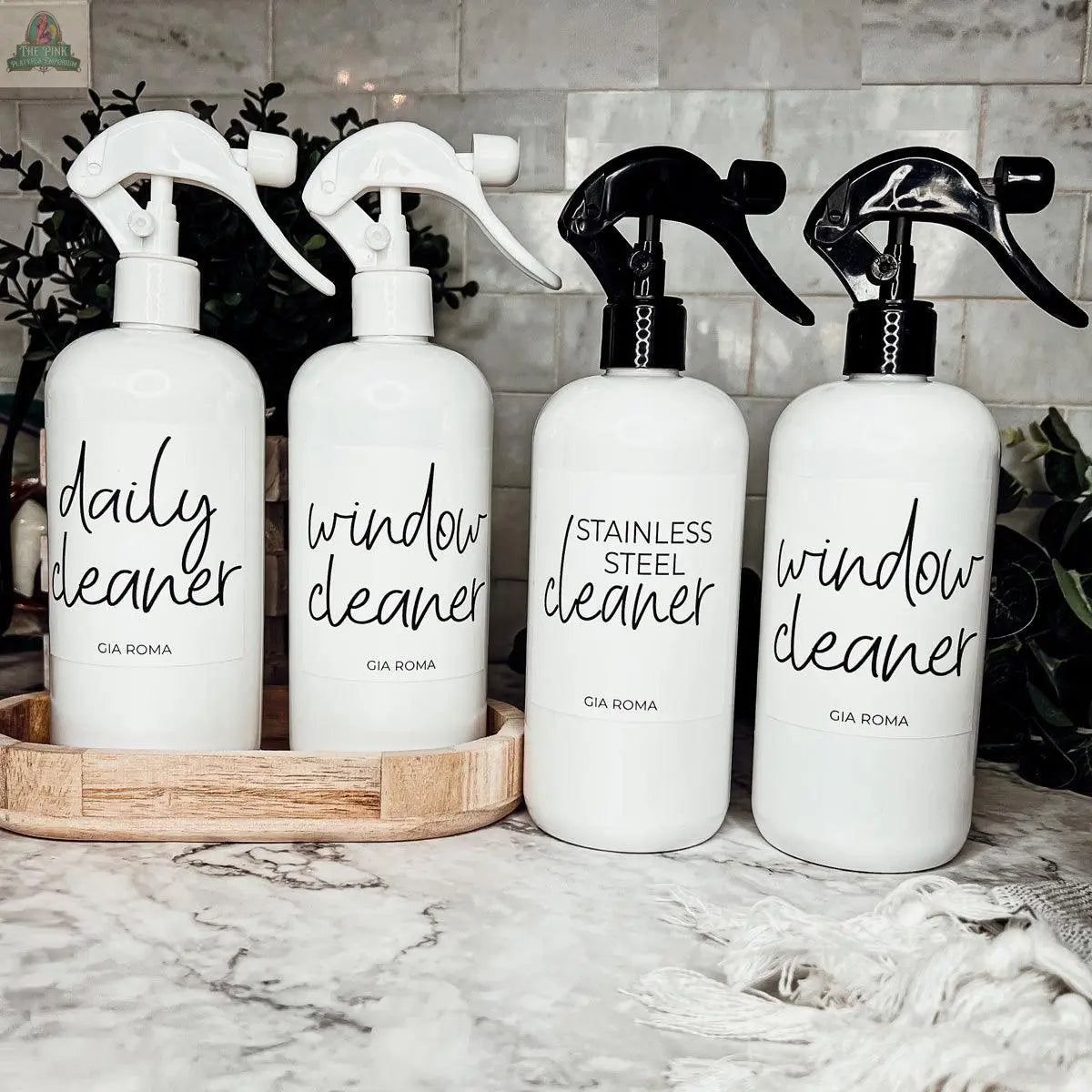 Four 16oz Plastic Cleaning Bottles with black or white spray pumps, labeled for daily, window, and stainless steel cleaner, sit on a marble countertop; two are in a wooden tray with greenery in the background.