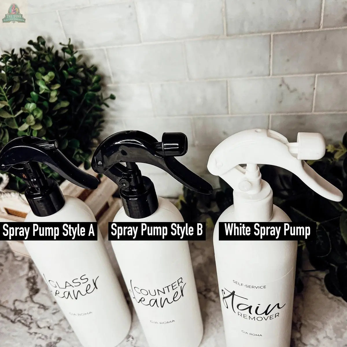 Three 16oz Plastic Cleaning Bottles with Style A, Style B, and White Spray Pump tops sit on a marble counter, surrounded by plants and a tiled backsplash. These reusable bottles are perfect for cleaning solutions.