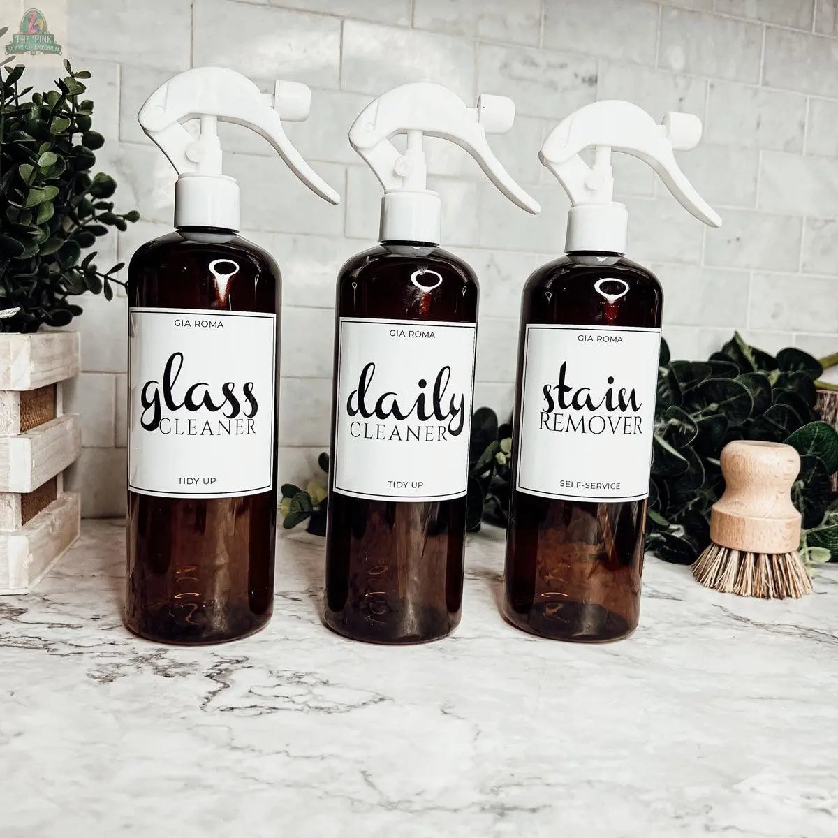 Three brown 16oz Plastic Cleaning Bottles labeled glass cleaner, daily cleaner, and stain remover stand on a marble countertop, with green plants and a wooden scrub brush in the background.