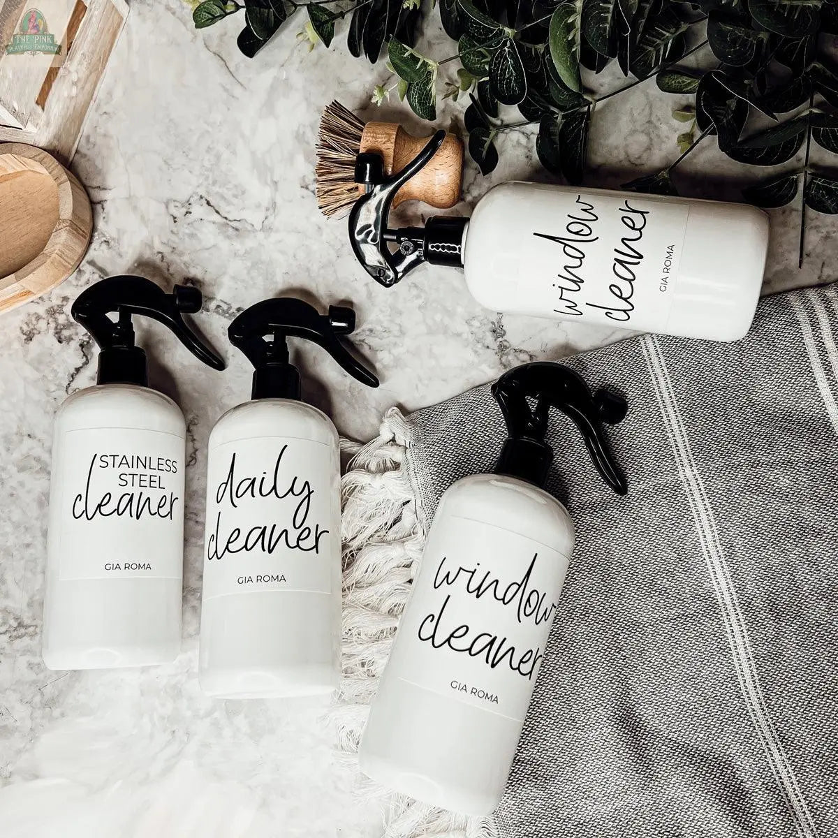 Four labeled 16oz Plastic Cleaning Bottles—stainless steel cleaner, daily cleaner, and two window cleaners—are arranged on a marble surface with a striped towel, brush, wooden tray, and green leaves, highlighting the stylish reusable dispensers.