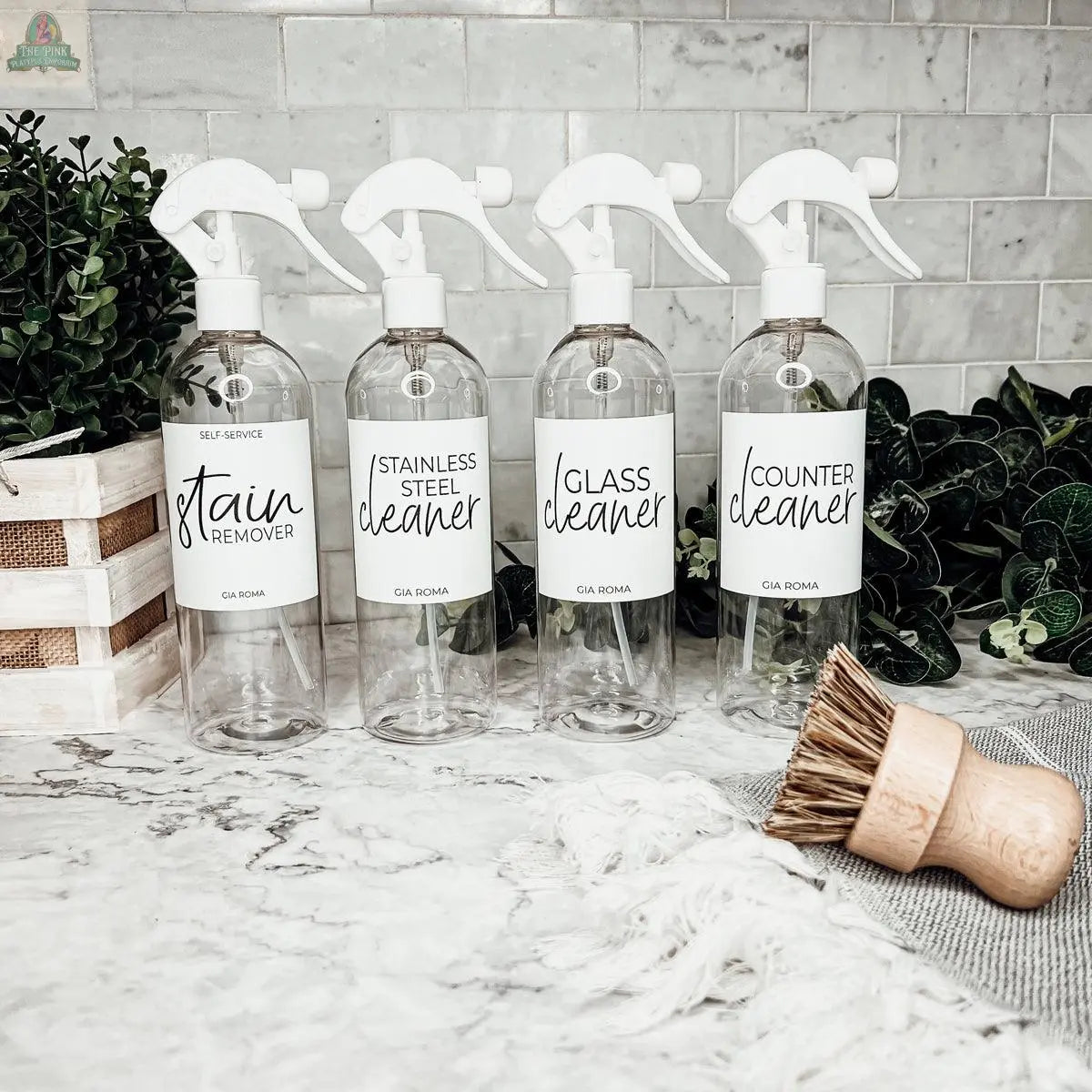 Four 16oz Plastic Cleaning Bottles, each labeled as stain remover, stainless steel cleaner, glass cleaner, and counter cleaner, are displayed on a marble countertop with plants and cleaning tools in the background as reusable dispensers.