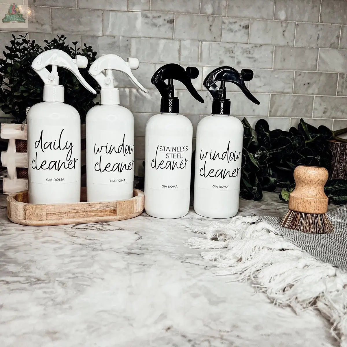 Four 16oz Plastic Cleaning Bottles, labeled daily, window, and stainless steel cleaner, stand on a counter with a scrub brush, greenery, and a beige cloth in a tiled kitchen or bathroom. Waterproof labels allow for easy reuse.