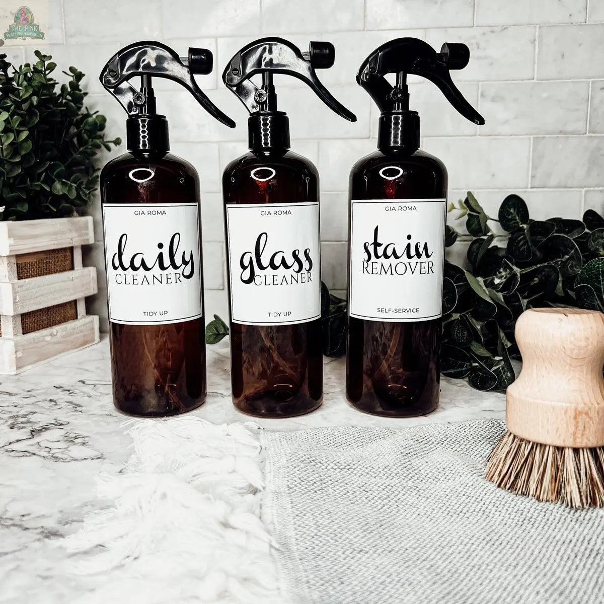 Three 16oz Plastic Cleaning Bottles with waterproof labels—Daily Cleaner, Glass Cleaner, and Stain Remover—sit on a marble countertop with greenery, a scrub brush, and a white cloth in the background.