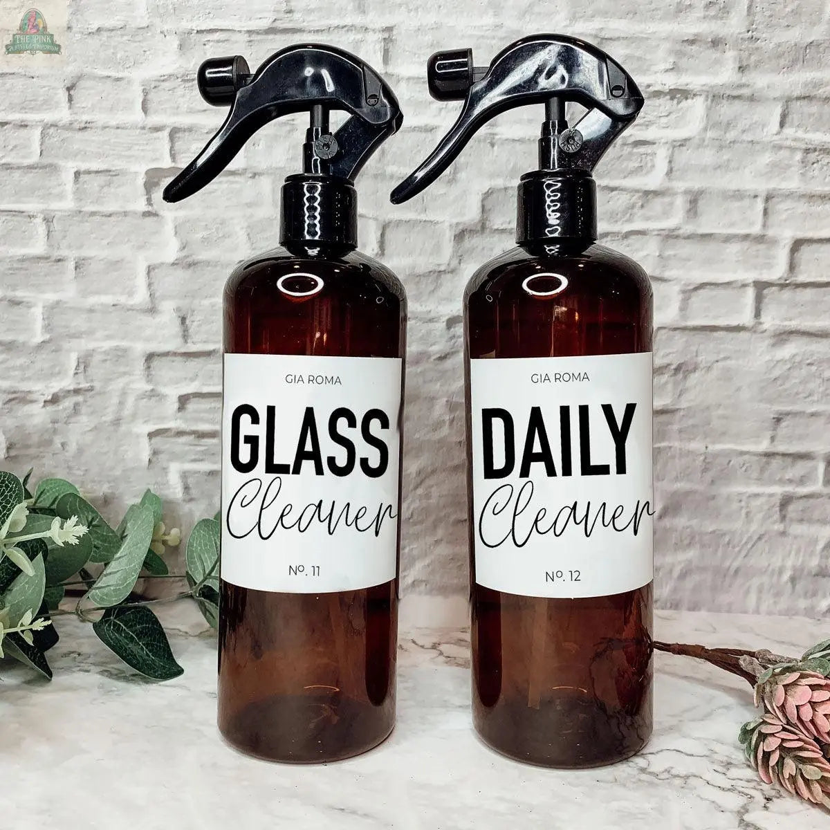 Two brown 16oz Plastic Cleaning Bottles with black spray nozzles, labeled Glass Cleaner and Daily Cleaner, feature waterproof labels and are displayed on a counter with a white textured brick background and green plants beside them.