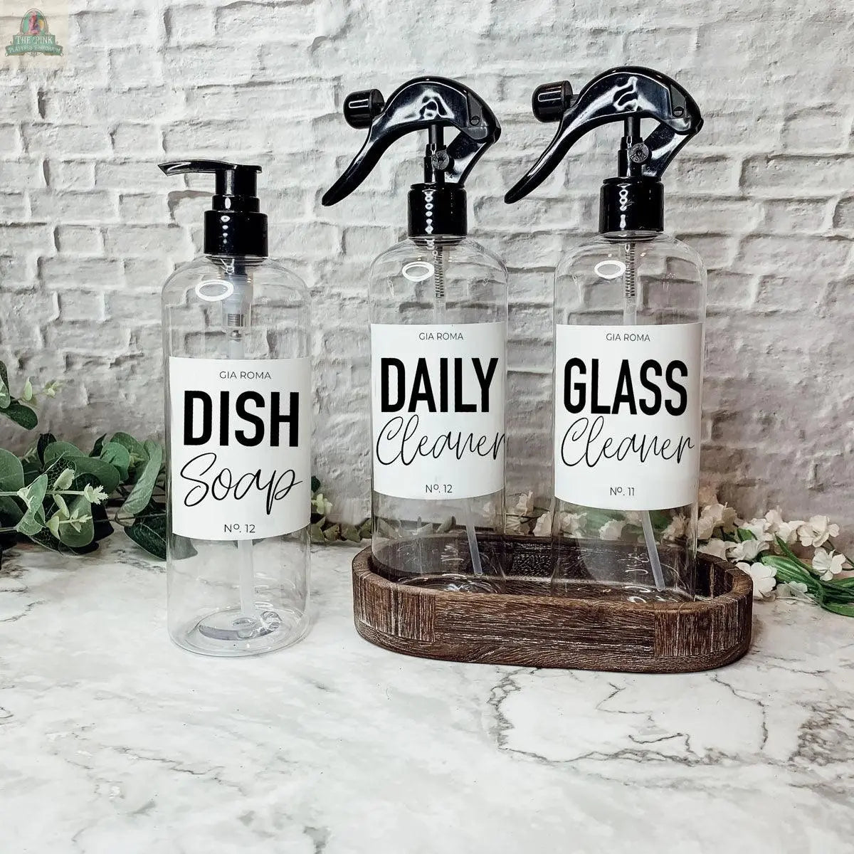 Three 16oz Plastic Cleaning Bottles—a pump for Dish Soap and two spray bottles for Daily Cleaner and Glass Cleaner—sit in a wooden tray on a marble counter, set against a white brick wall.