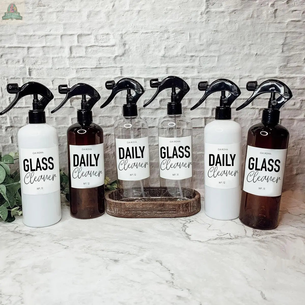 Six 16oz Plastic Cleaning Bottles—labeled GLASS Cleaner and DAILY Cleaner—are arranged on a marble countertop with a small tray and a green plant, set against a white brick background.