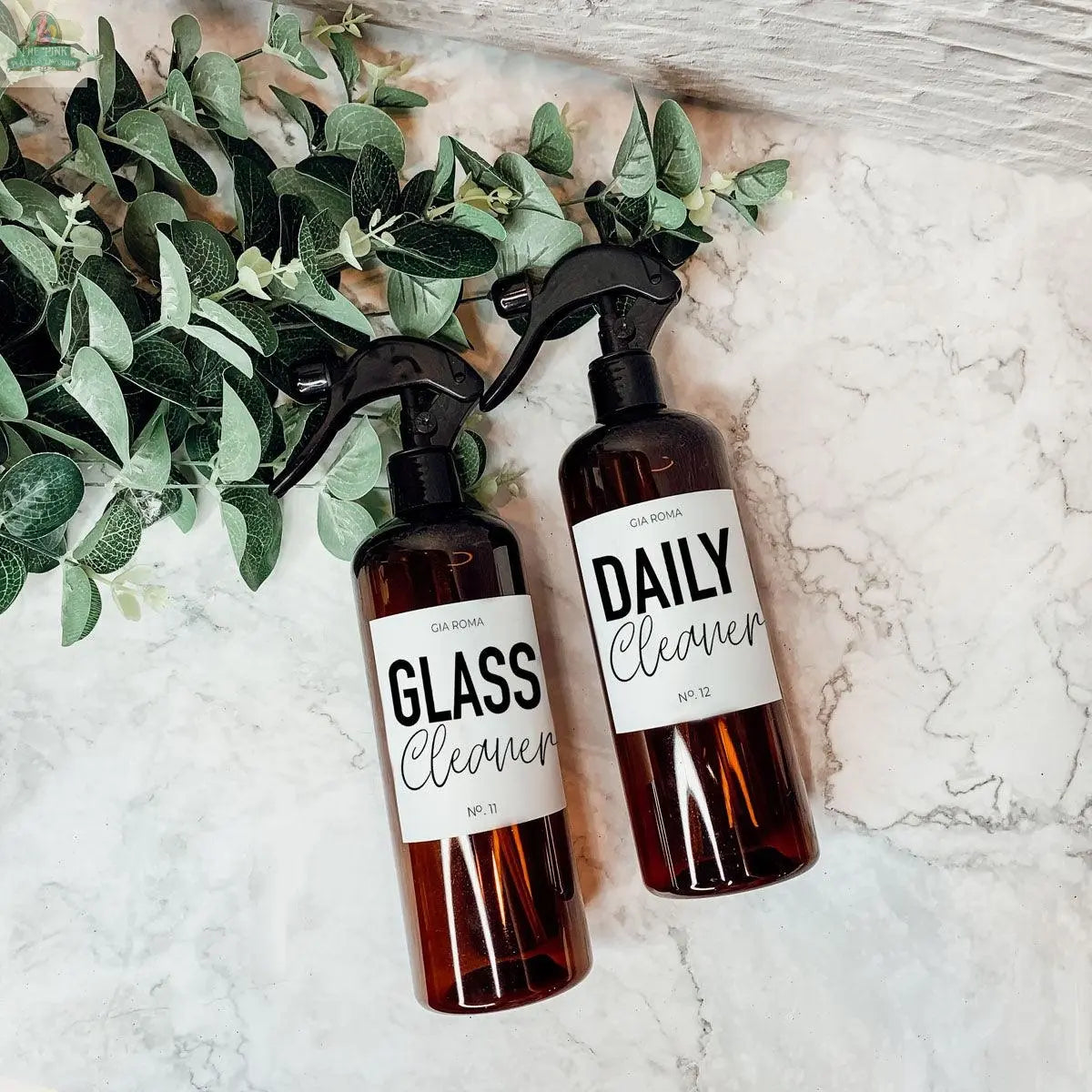 Two 16oz Plastic Cleaning Bottles with black spray nozzles, labeled GLASS Cleaner and DAILY Cleaner, rest on a marble surface beside green leaves—stylish and reusable dispensers for your daily cleaning needs.