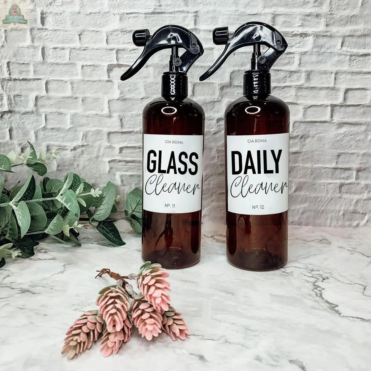 Two 16oz Plastic Cleaning Bottles labeled Glass Cleaner and Daily Cleaner stand on a marble surface with green and pink artificial plants, set against a white brick background.
