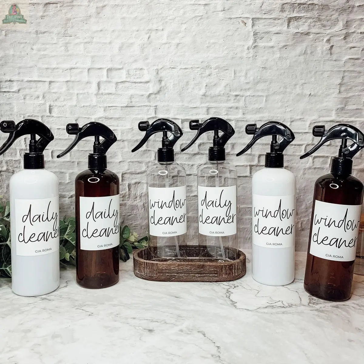 Six 16oz Plastic Cleaning Bottles—labeled Daily Cleaner and Window Cleaner, in brown, white, and clear with waterproof labels and spray tops—are displayed on a counter before a white brick wall with greenery in the background.