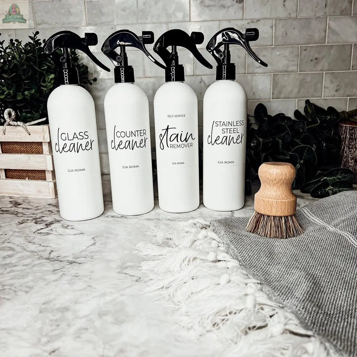 Four 16oz Plastic Cleaning Bottles labeled Glass Cleaner, Counter Cleaner, Stain Remover, and Stainless Steel Cleaner with spray tops stand on a marble countertop, surrounded by a scrub brush, towel, and green plants.