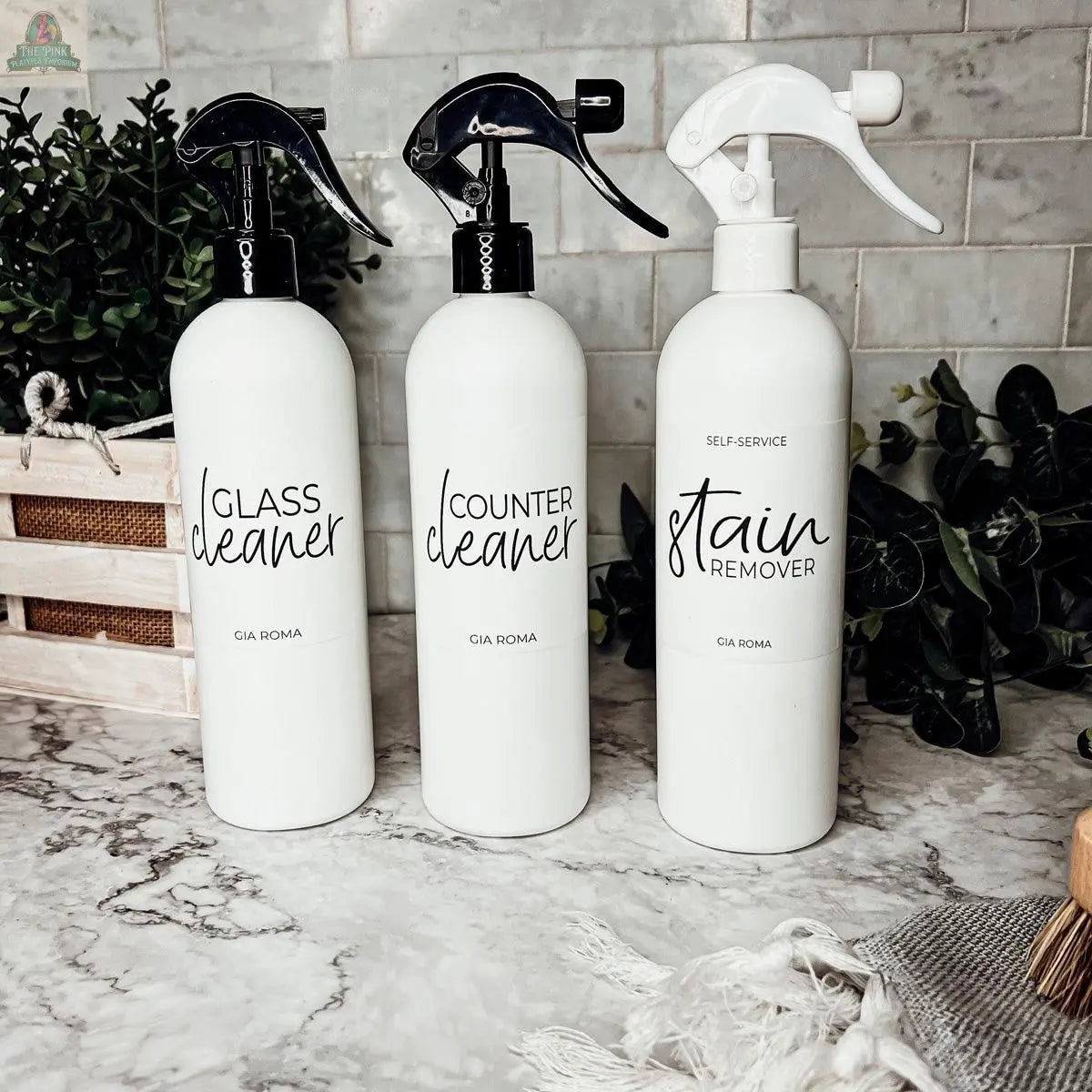 Three 16oz Plastic Cleaning Bottles with waterproof labels—Glass Cleaner, Counter Cleaner, and Stain Remover—sit on a marble countertop, with green plants and a wooden tray in the background.