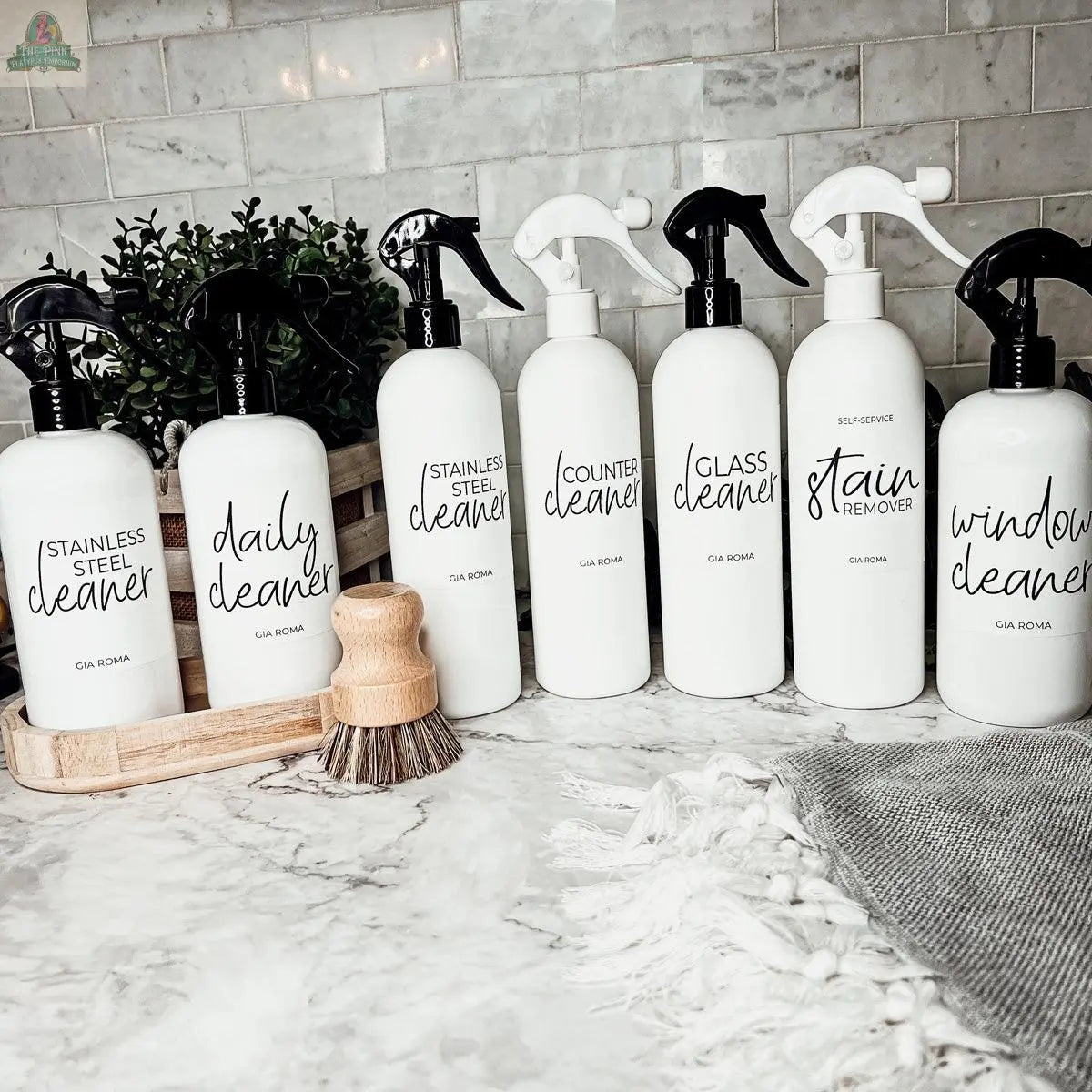 Six 16oz Plastic Cleaning Bottles with black and white spray pump nozzles, each labeled for different cleaners, are arranged on a marble counter beside a scrub brush, with a small potted plant and subway tile backsplash in the background.