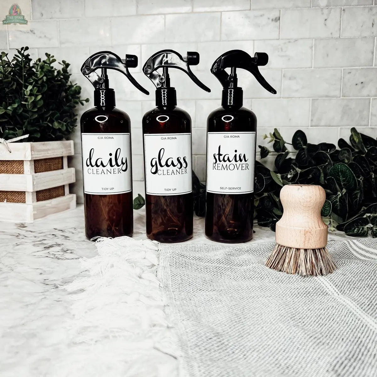 Three 16oz Plastic Cleaning Bottles labeled “daily cleaner,” “glass cleaner,” and “stain remover” sit on a marble counter with a scrub brush, greenery, and a linen towel, set against a tiled backsplash.