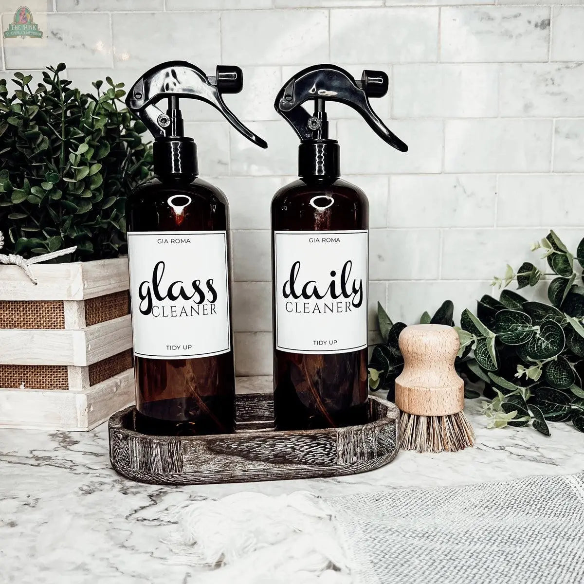 Two 16oz Plastic Cleaning Bottles labeled glass cleaner and daily cleaner sit in a wooden tray on a marble countertop, surrounded by green plants, a wooden scrub brush, and a white tile backsplash.