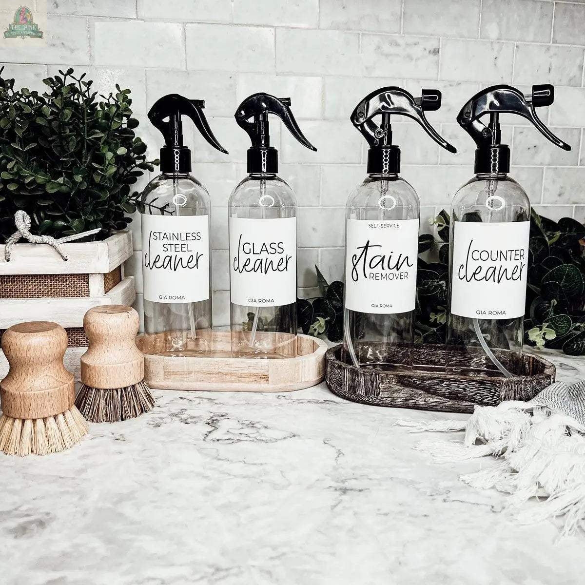 Four 16oz Plastic Cleaning Bottles—Stainless Steel Cleaner, Glass Cleaner, Stain Remover, Counter Cleaner—each with spray pump and waterproof label, are displayed on a marble counter with brushes, trays, and green plants in the background.