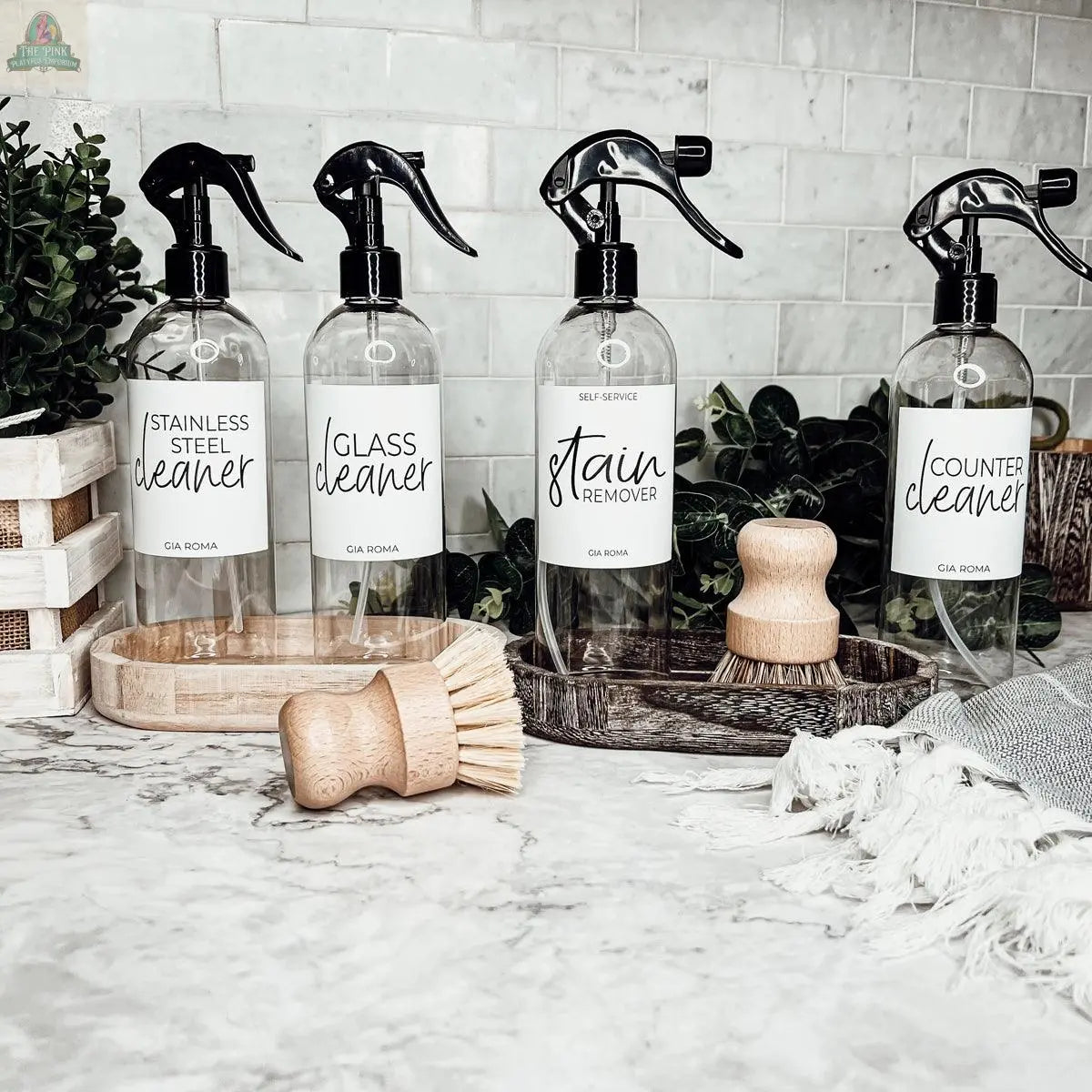 Four 16oz Plastic Cleaning Bottles—labeled stainless steel cleaner, glass cleaner, stain remover, and counter cleaner—sit on a marble countertop with brushes, cloth, and greenery in the background. Each has a convenient spray pump design.