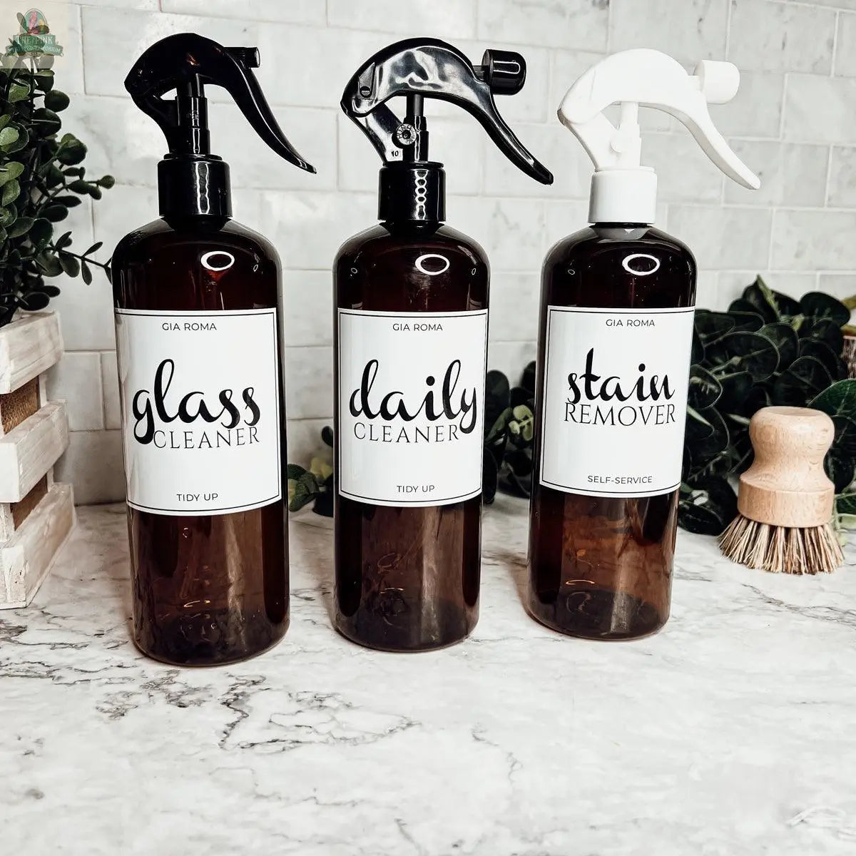 Three 16oz Plastic Cleaning Bottles, labeled glass cleaner, daily cleaner, and stain remover with waterproof labels, stand on a marble countertop among plants and cleaning brushes.
