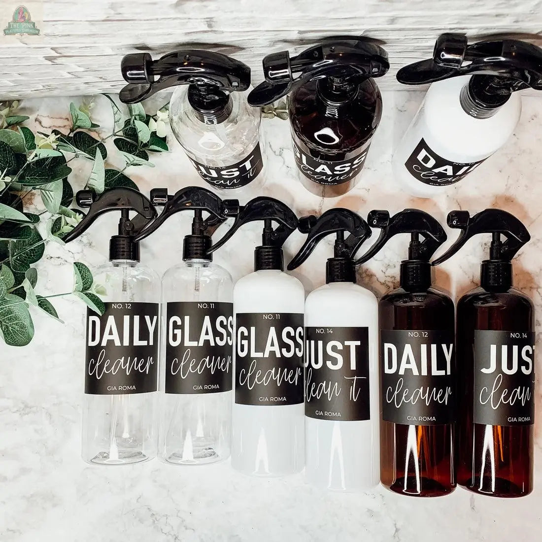 A row of 16oz Plastic Cleaning Bottles with waterproof labels (DAILY cleaner, GLASS cleaner, JUST clean it) sits on marble beside green leafy plants—ideal reusable dispensers to upgrade your cleaning routine.