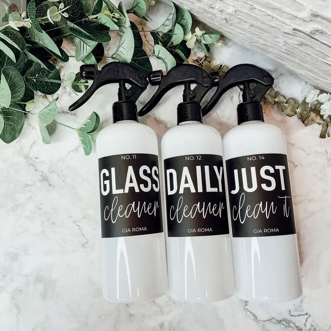Three 16oz Plastic Cleaning Bottles with black waterproof labels—GLASS cleaner, DAILY cleaner, and JUST clean it—are arranged on a marble surface with green leafy branches nearby.