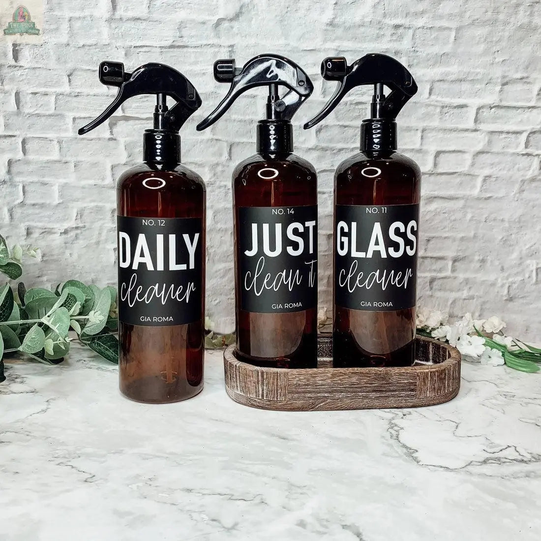Three brown 16oz Plastic Cleaning Bottles with waterproof labels—Daily Cleaner, Just Clean It, and Glass Cleaner—sit on a white marble surface with a wooden tray, against a white brick background and green foliage.