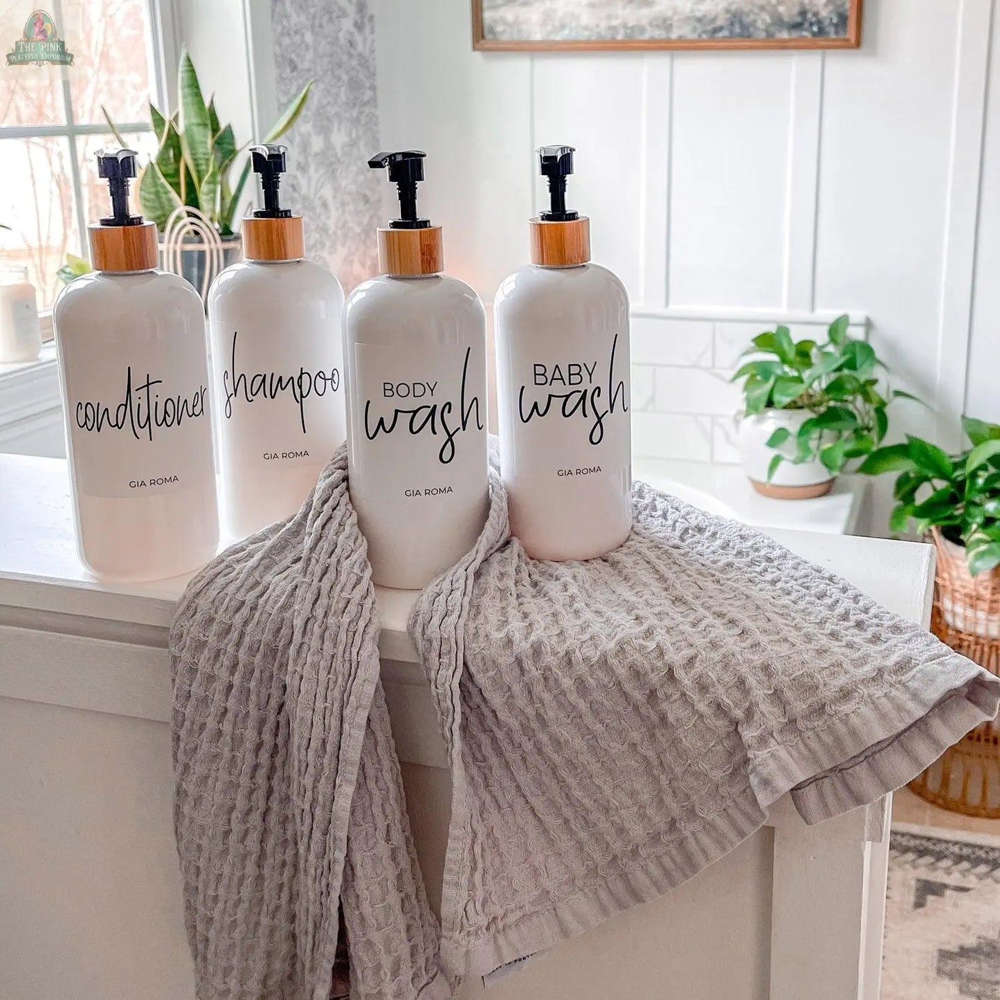 Four 16oz Plastic White | White Label bottles—conditioner, shampoo, body wash, and baby wash—sit on a white surface with two gray towels. Each refillable dispenser has a waterproof label, with green houseplants and natural light in the background.