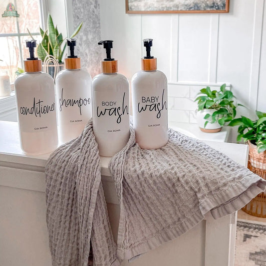 Four 16oz Plastic White | White Label bottles—conditioner, shampoo, body wash, and baby wash—sit on a white surface with two gray towels. Each refillable dispenser has a waterproof label, with green houseplants and natural light in the background.