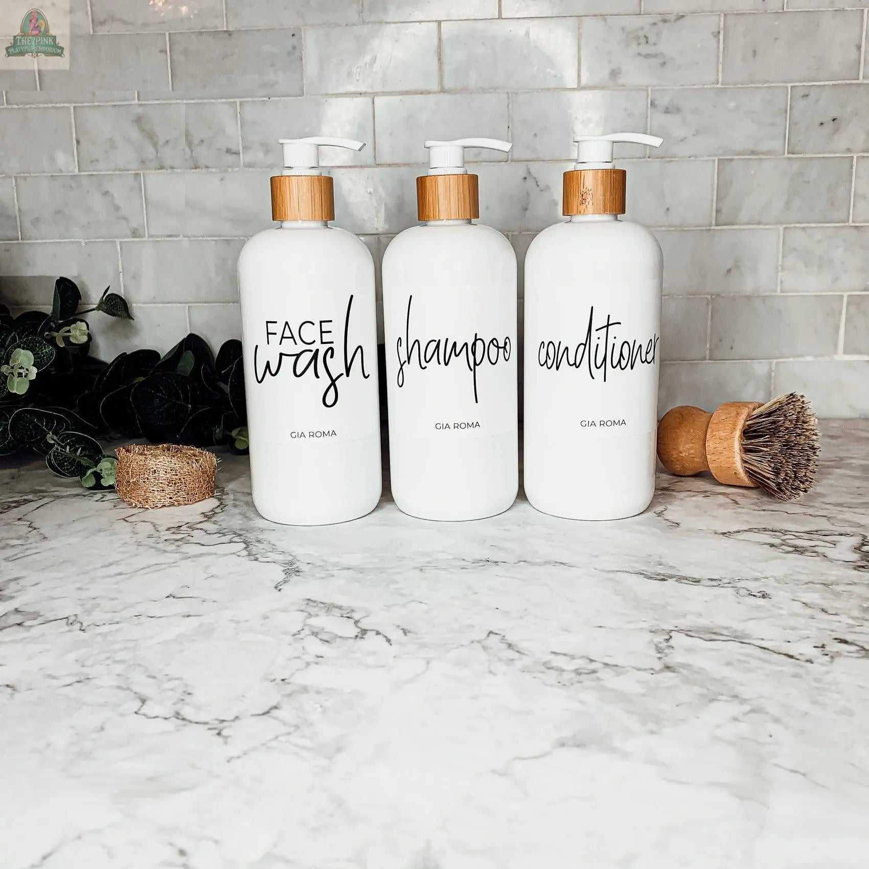 Three 16oz Plastic White | White Label bottles for Face Wash, Shampoo, and Conditioner sit on a marble countertop with a tile backsplash. These refillable dispensers have waterproof labels and are accented by a plant, brush, and sponge.