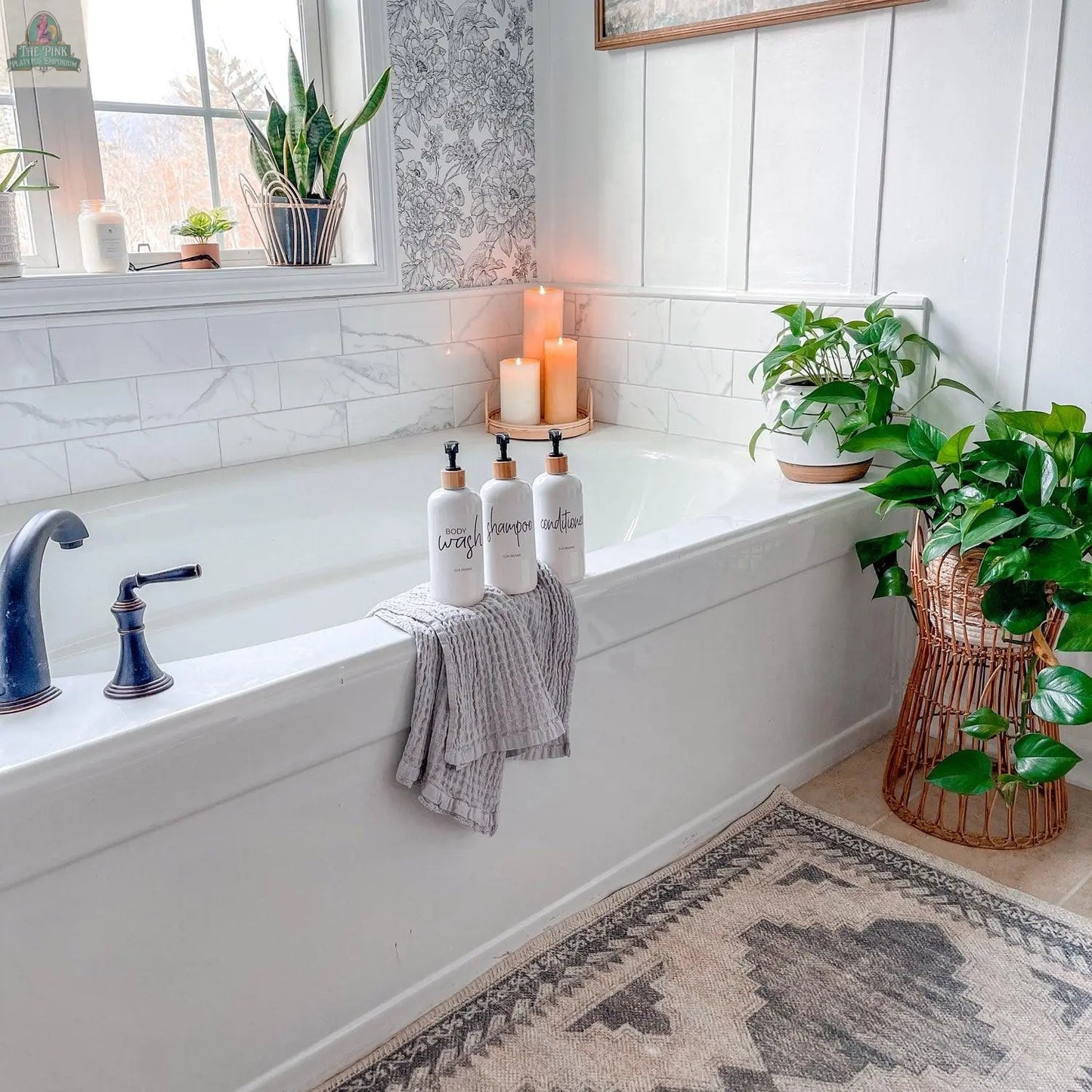 A bright, modern bathroom features a white bathtub, black faucet, gray striped towel, three 16oz Plastic White | White Label dispensers, lit candles, potted green plants, and a patterned rug on the floor.