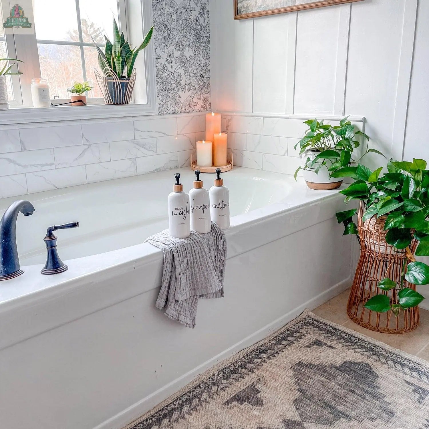 A bright, modern bathroom features a white bathtub, black faucet, gray striped towel, three 16oz Plastic White | White Label dispensers, lit candles, potted green plants, and a patterned rug on the floor.