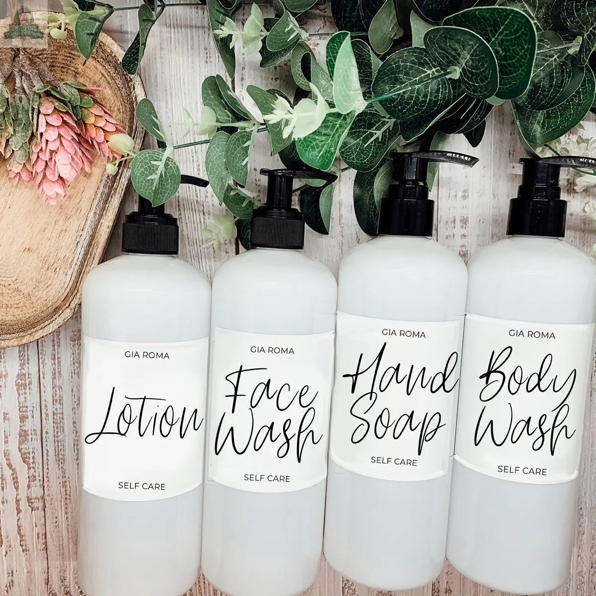 Four 16oz Plastic White | White Label bottles—labeled Lotion, Face Wash, Hand Soap, and Body Wash—stand in a row on wood with greenery and pink flowers nearby. The refillable dispensers have waterproof labels for lasting style.
