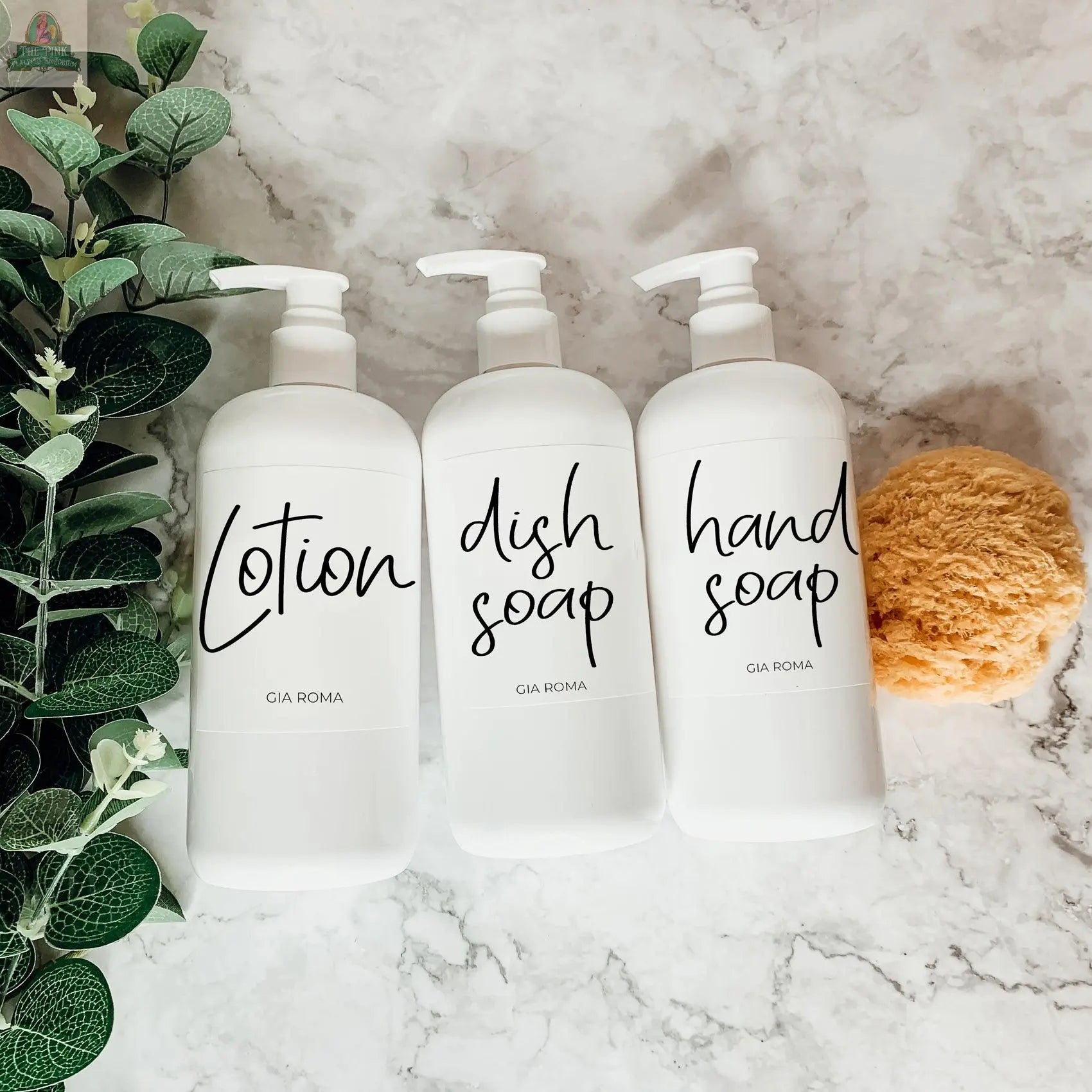 Three 16oz Plastic White | White Label bottles are arranged on a marble surface near a leafy plant and a yellow sponge. These refillable dispensers, labeled for lotion, dish soap, and hand soap, bring elegance to any space.