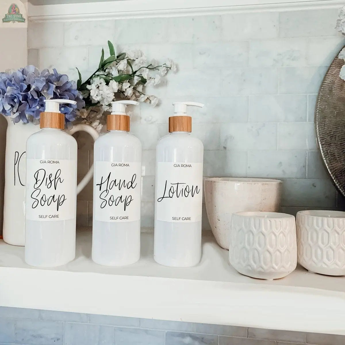 Three 16oz Plastic White | White Label bottles labeled “Dish Soap,” “Hand Soap,” and “Lotion” are arranged on a white shelf amid textured cups and purple and white flowers, creating a bright, clean atmosphere.