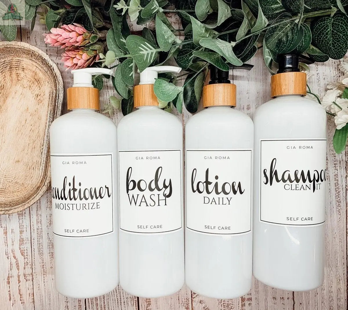 Four 16oz Plastic White bottles with white waterproof labels—ideal for Conditioner, Body Wash, Lotion, and Shampoo—sit side by side on a wooden tray surrounded by greenery, serving as stylish, refillable dispensers.