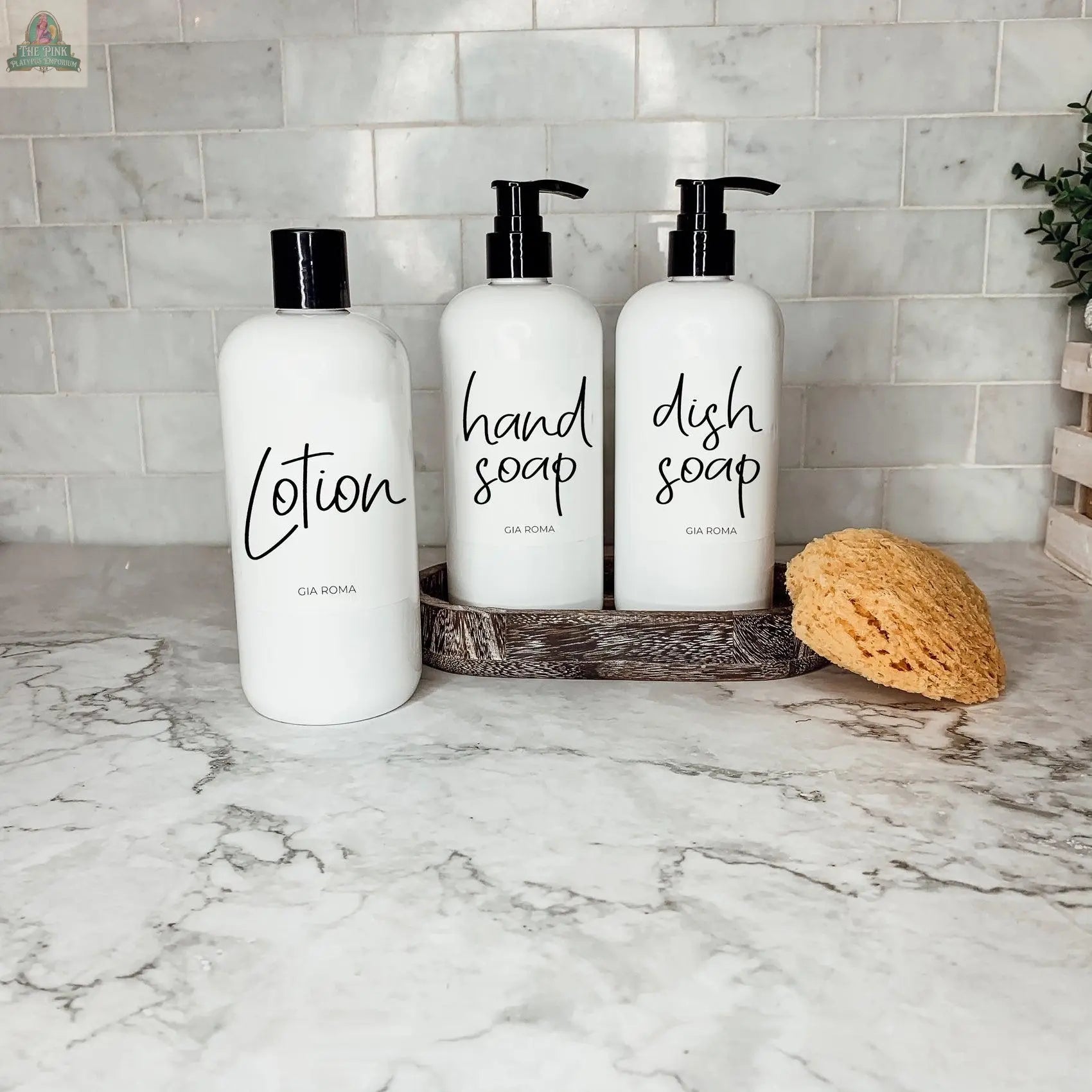 Three 16oz Plastic White | White Label bottles—lotion, hand soap, and dish soap—sit in a tray on a marble countertop. A yellow sponge rests beside the refillable dispensers, with a tiled backsplash in the background.