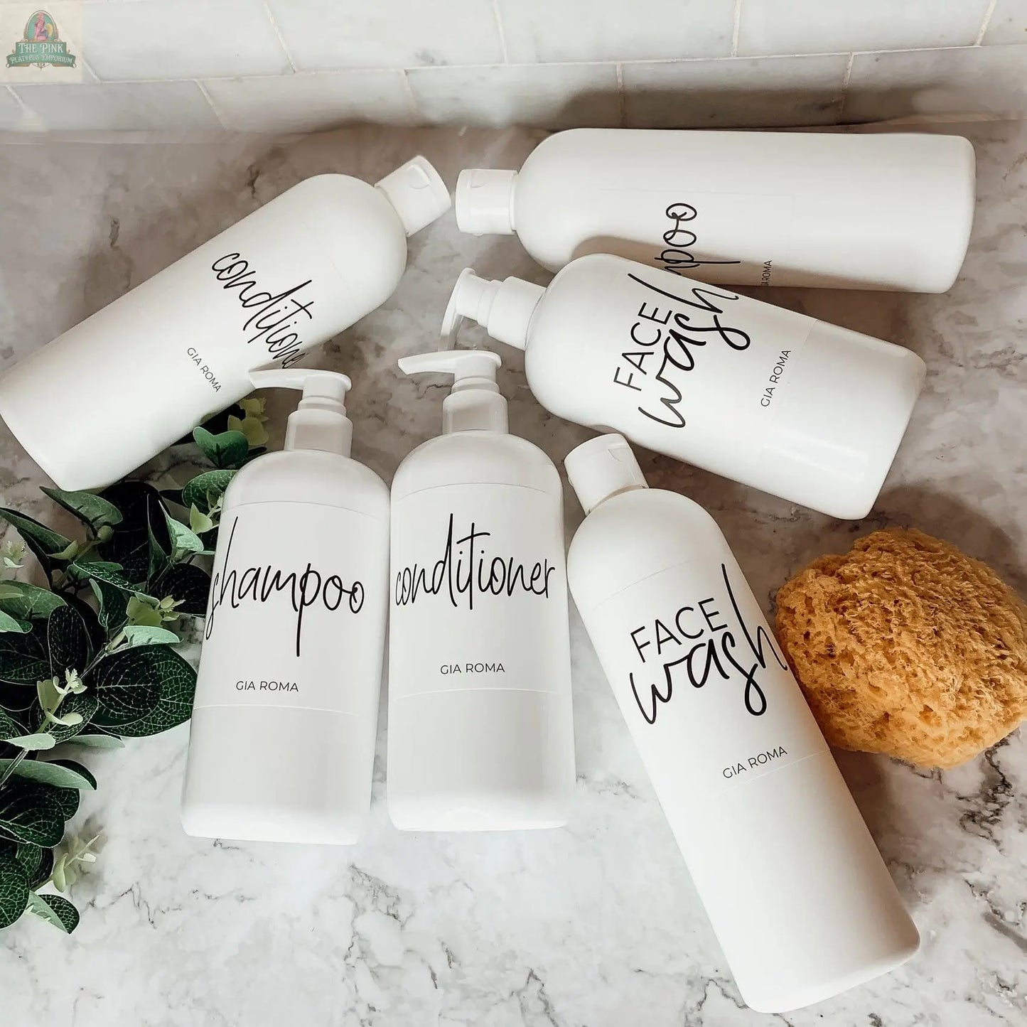 A marble countertop displays 16oz Plastic White | White Label bottles for shampoo, conditioner, and face wash. Some dispensers have pumps; others lie flat. A natural sponge and a green plant are also present.