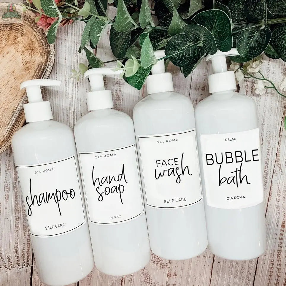Four 16oz Plastic White | White Label bottles labeled shampoo, hand soap, face wash, and bubble bath sit side by side on a wooden surface, accented by nearby greenery and a wooden tray; these dispensers are refillable.