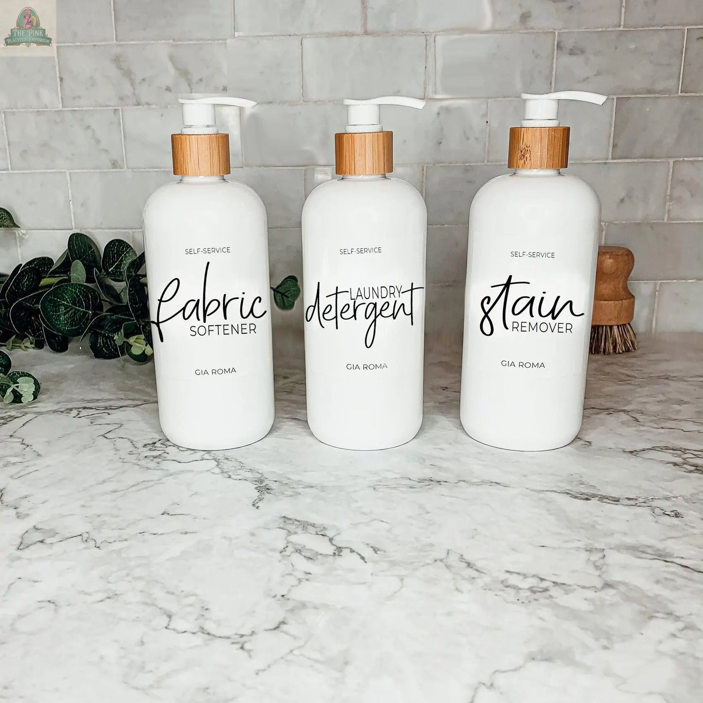 Three 16oz Plastic White bottles with white, waterproof labels—labeled Fabric Softener, Laundry Detergent, and Stain Remover—sit on a marble countertop. These practical, stylish dispensers are refillable for everyday laundry use.