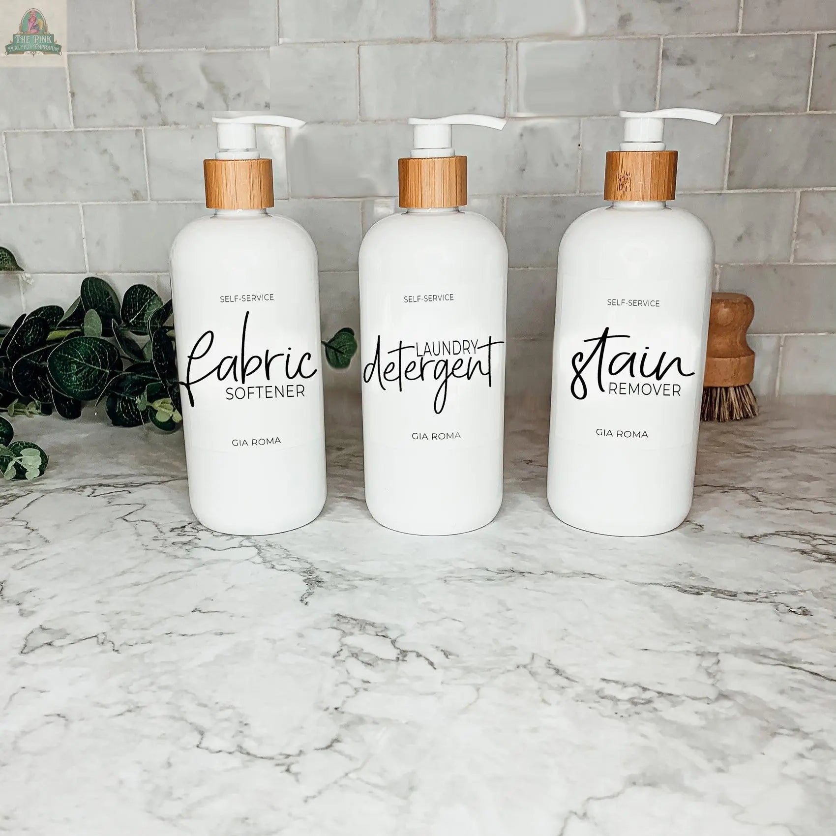 Three 16oz Plastic White bottles with white, waterproof labels—labeled Fabric Softener, Laundry Detergent, and Stain Remover—sit on a marble countertop. These practical, stylish dispensers are refillable for everyday laundry use.
