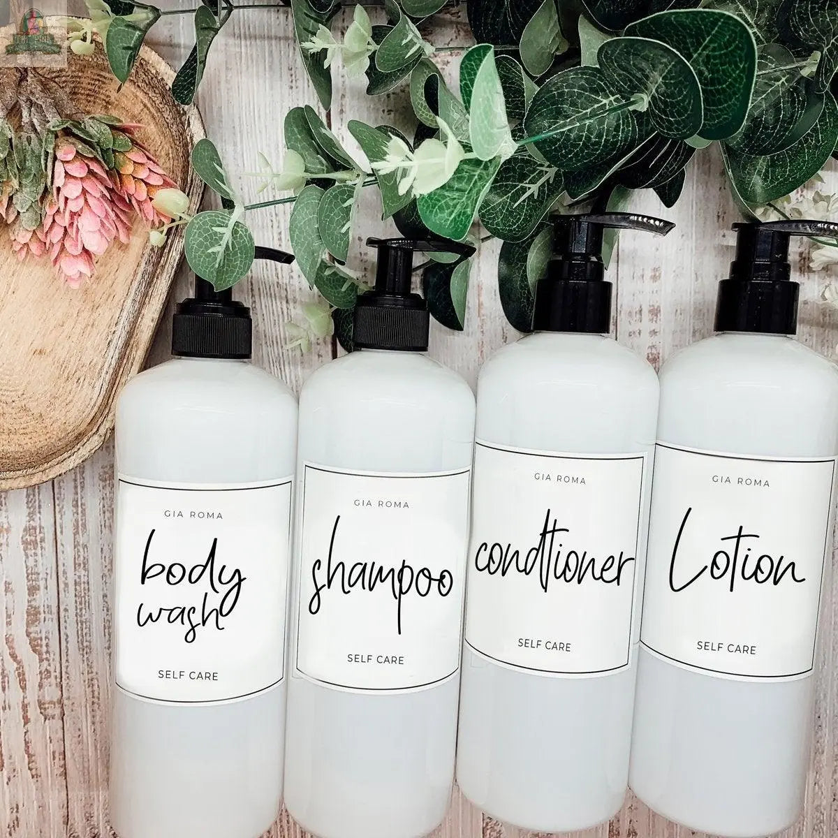 Four 16oz Plastic White bottles with white labels stand in a row on a rustic wooden surface. Each refillable dispenser is labeled for body wash, shampoo, conditioner, and lotion, with waterproof labels, green leaves, and flowers above.