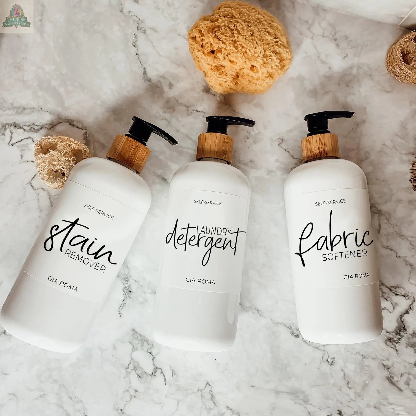 Three 16oz Plastic White bottles with White Label—filled with Stain Remover, Laundry Detergent, and Fabric Softener—serve as chic refillable dispensers on a marble surface, accented by natural sponges.