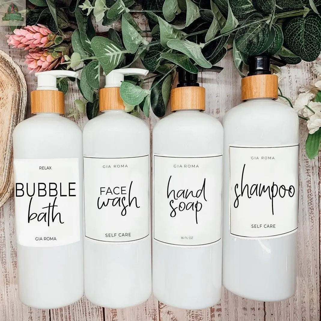 Four 16oz Plastic White | White Label refillable dispensers—labeled bubble bath, face wash, hand soap, and shampoo—are lined up in a row with waterproof labels and bamboo lids, greenery in the background.