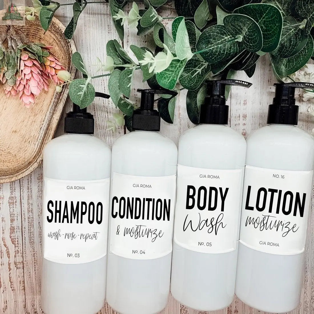Four 16oz Plastic White | White Label bottles, labeled shampoo, conditioner, body wash, and lotion, are arranged on a wood surface with green leaves and a pink flower. Clear, minimalist waterproof labels make them ideal refillable dispensers.