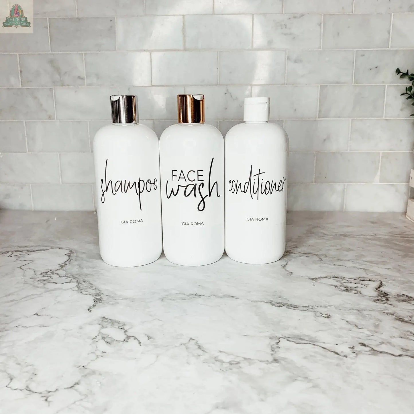 Three 16oz Plastic White | White Label bottles labeled shampoo, face wash, and conditioner sit on a marble countertop with a white subway tile backsplash. Each refillable dispenser features simple black text and either metallic or white pump tops.