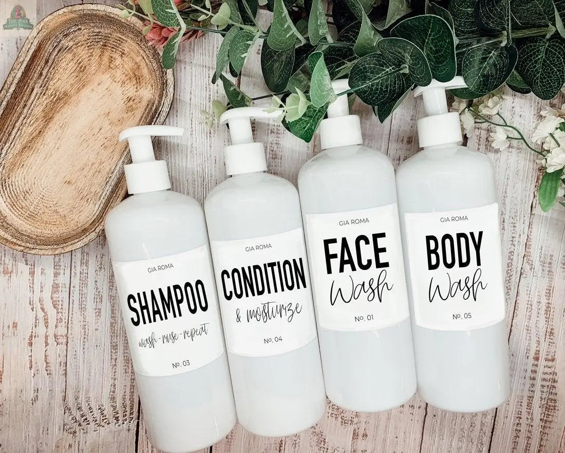Four 16oz Plastic White bottles with durable white labels—Shampoo, Condition & Moisturize, Face Wash, and Body Wash—are set up as refillable dispensers on a wooden surface, with greenery and a wooden tray beside them.