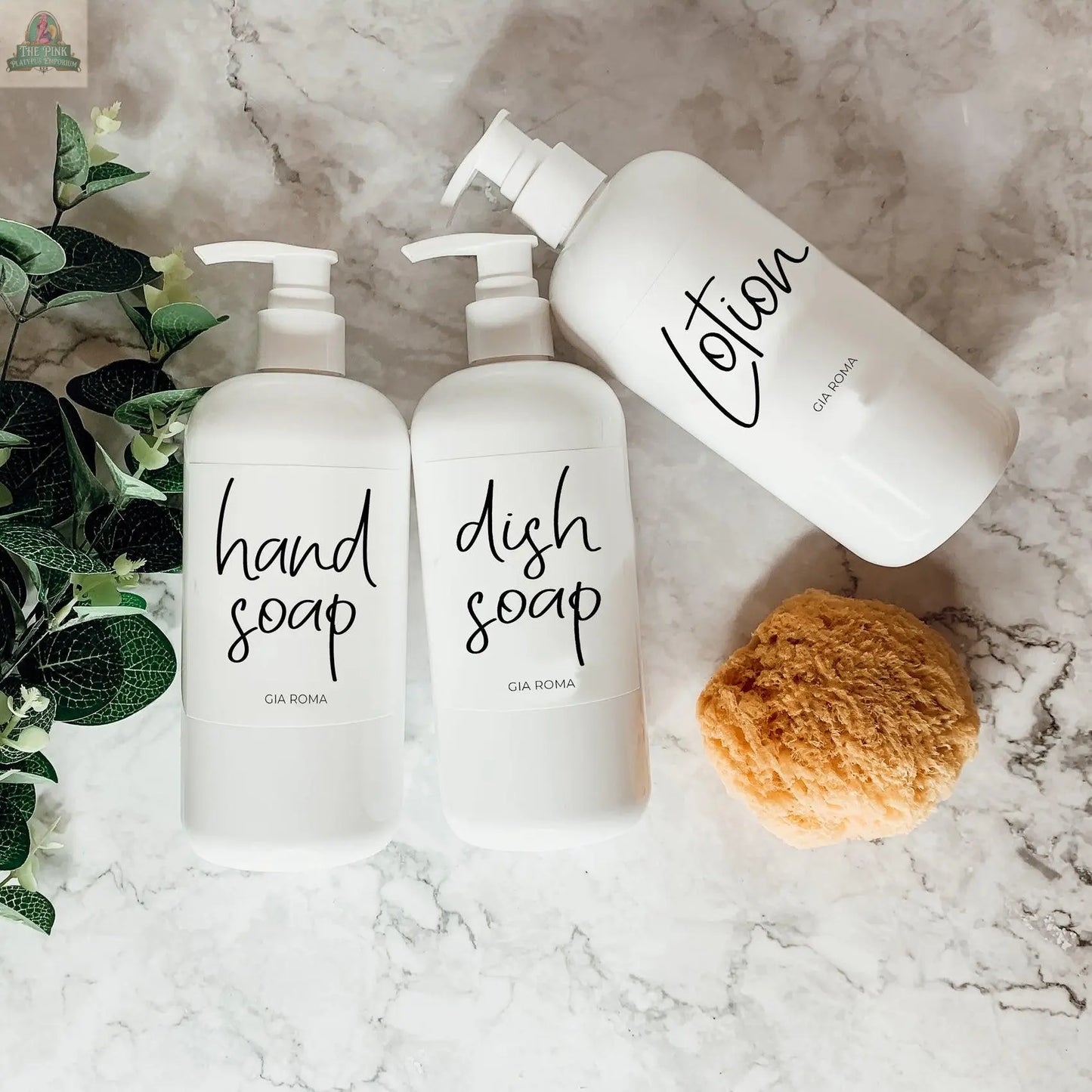 Three 16oz Plastic White | White Label bottles labeled hand soap, dish soap, and lotion sit on a marble surface beside a yellow sponge and green leaves. These refillable dispensers have modern, handwritten-style text and waterproof labels.