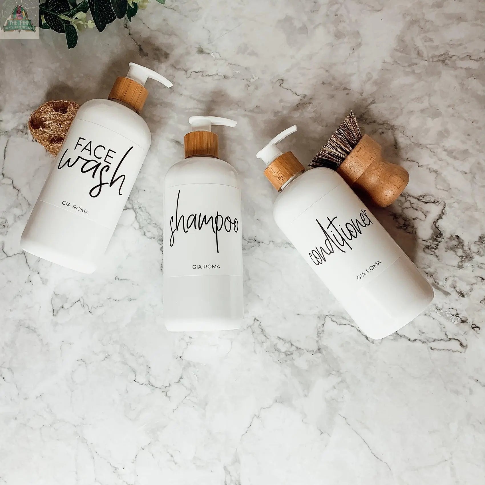 Three 16oz Plastic White | White Label bottles labeled Face Wash, Shampoo, and Conditioner rest on a marble surface, with a wooden brush, sponge, and green leaves partially visible around them. Each bottle features a waterproof label.