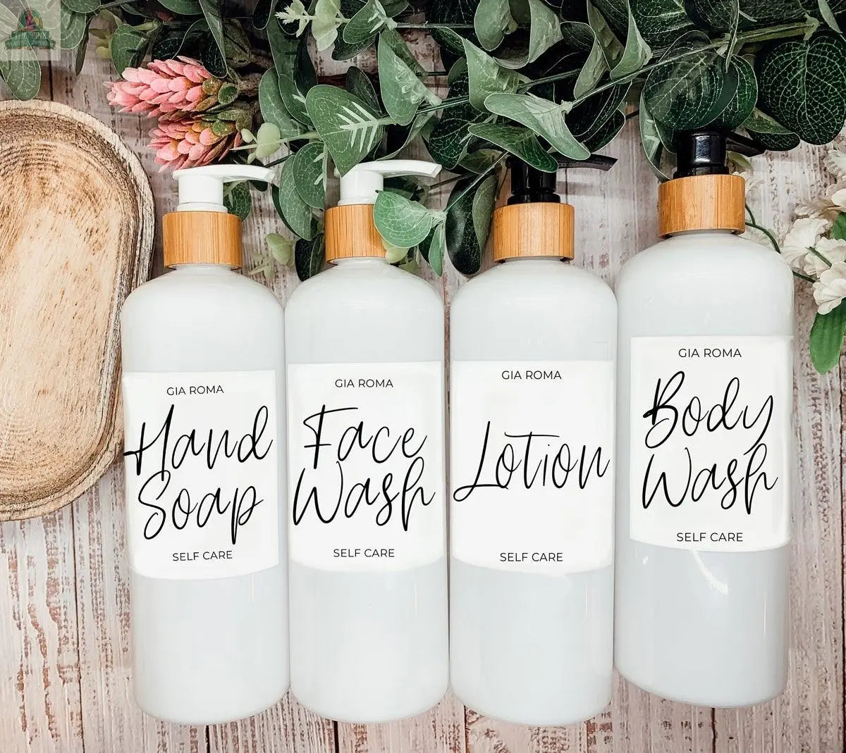 Four 16oz Plastic White | White Label bottles with wooden pumps—dispensing Hand Soap, Face Wash, Lotion, and Body Wash—stand stylishly refillable on a wooden surface, accented by green leaves and a wooden tray.