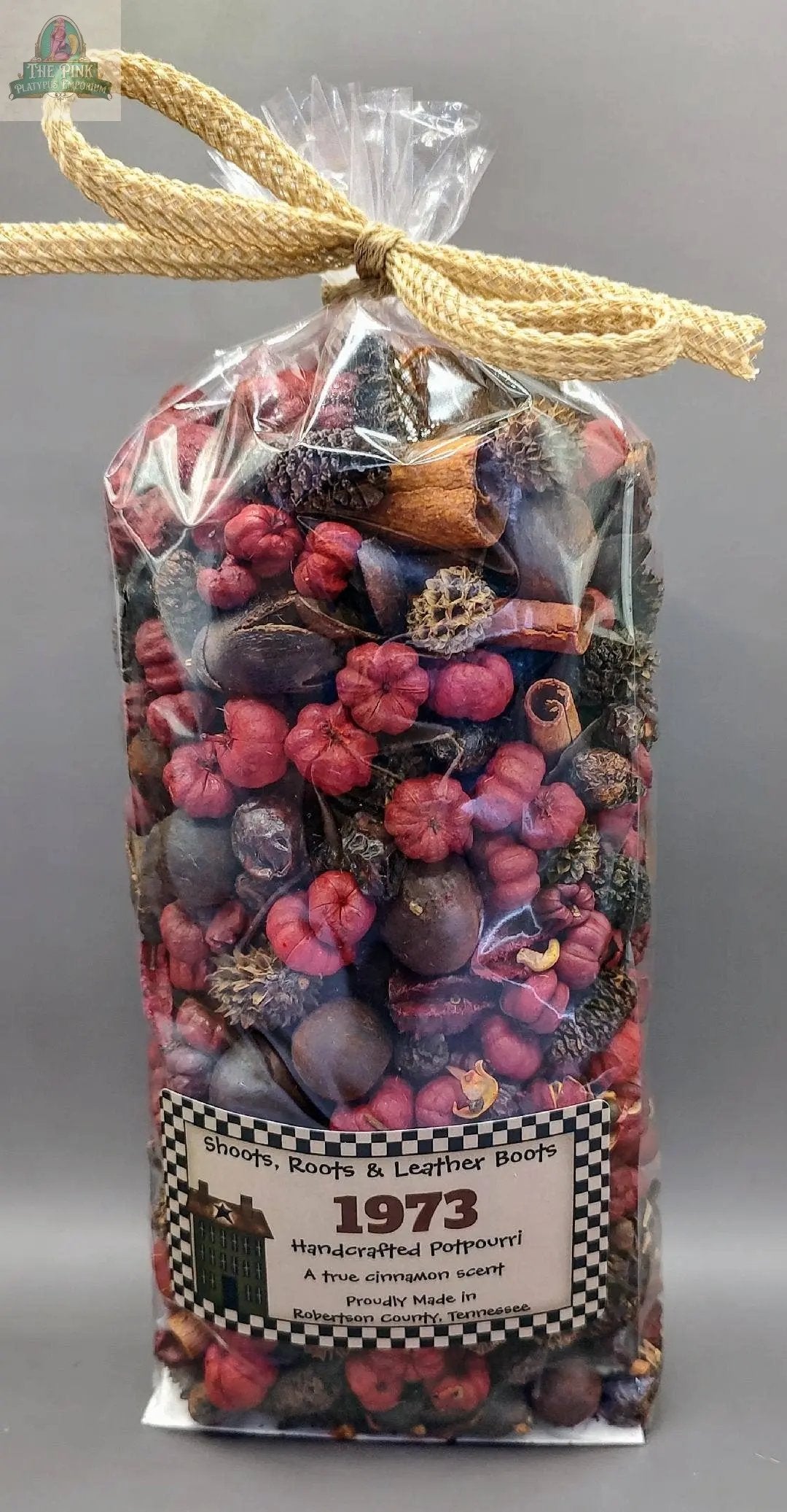 A clear bag tied with tan rope contains colorful, highly scented "1973" (A True Cinnamon Scent) Handcrafted Potpourri with red pods, pinecones, and cinnamon sticks. Label: “Shoots, Roots & Leather Boots 1973 Handcrafted Potpourri.”.