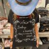 Person wearing a cowboy hat and black t-shirt with white text saying "darling a beautiful thing is never perfect" 