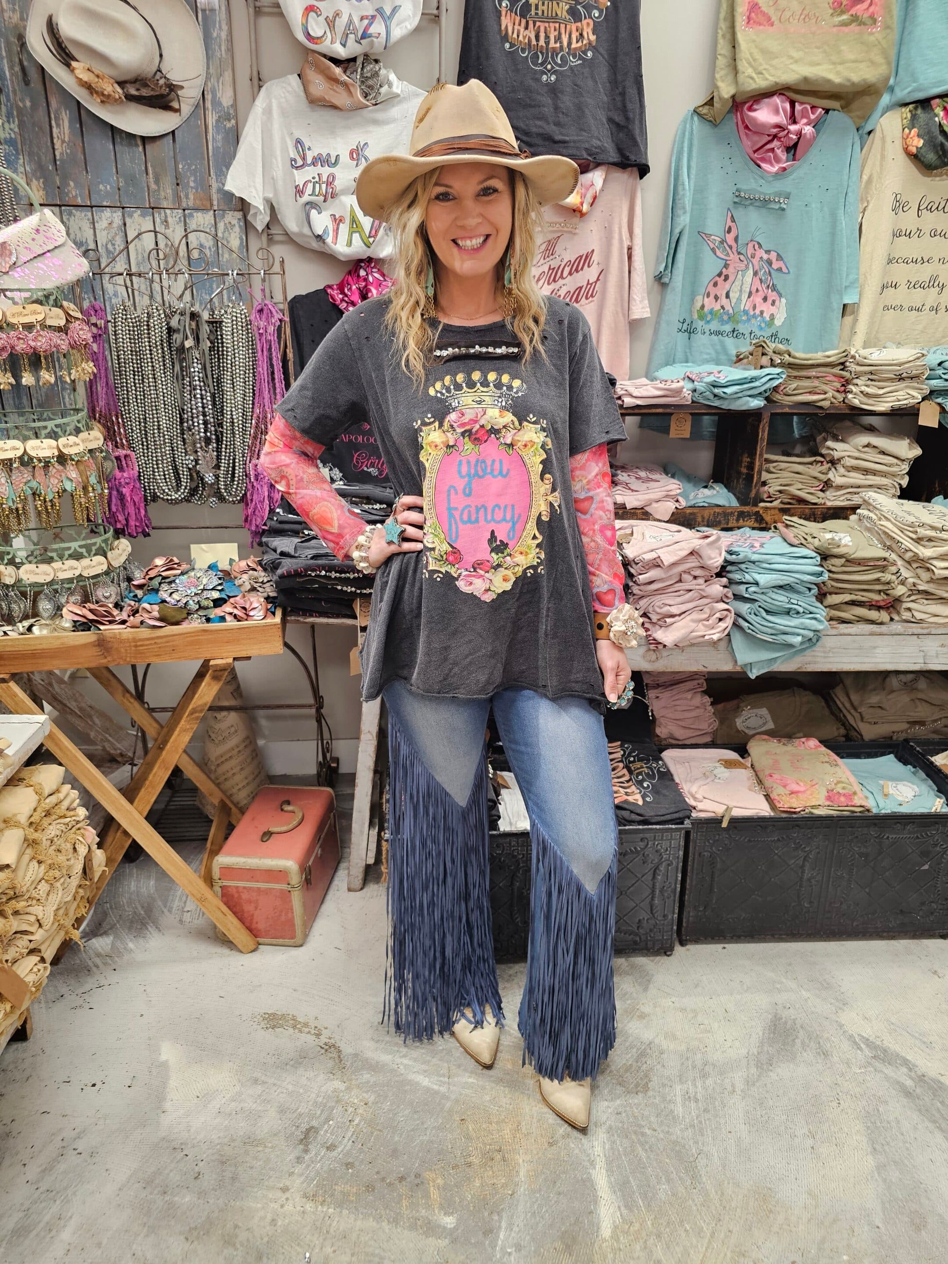 Woman wearing fringe jeans and cowboy hat in a store with various items on display and a graphic tshirt with the text "you fancy"